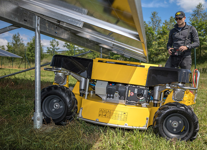 Ferngesteuerter Großflächenmäher Spider 2SGS Super Low beim Mähen unter einer niedrigen Photovoltaikanlage, im Hintergrund steht der Bediener mit der Fernsteuerung.