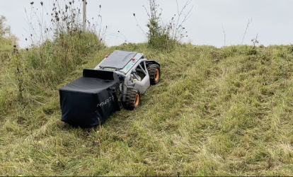 Das Bild zeigt den Mowrator im Einsatz auf einer starken Steigung mit unebenem Untergrund. Der Mäher wird von hinten links fotografiert und hat einen großen, schwarzen Grasfangsack aus Gewebe montiert. Er fährt eine anspruchsvolle, hügelige Wiesenfläche mit naturbelassenem Gras hinauf, was die Geländegängigkeit und Kraft des Geräts demonstriert.