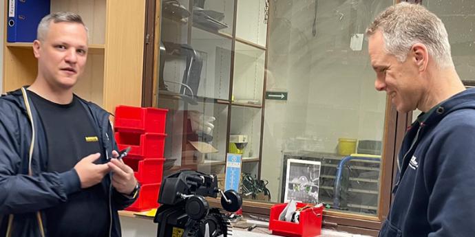Eine Szene in einem technischen Servicebereich oder Werkstatt. Zwei Personen in dunkler Arbeitskleidung stehen sich gegenüber und scheinen ein technisches Gerät oder Teil zu besprechen. Im Hintergrund sind Glasvitrinen mit Ausrüstung zu sehen, sowie rote Aufbewahrungsboxen. Die Umgebung wirkt professionell und organisiert, typisch für einen technischen Support- oder Wartungsbereich.