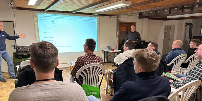 Ein Schulungsraum mit hölzerner Deckenverkleidung und Parkettboden. An der Wand ist eine Präsentation projiziert, die den Text 'Schulung Tag 1' zeigt. Im Raum sitzen mehrere Teilnehmer in weißen Gartenstühlen, die in Reihen angeordnet sind. Vorne links steht ein Referent, der mit einer einladenden Gestik etwas erklärt, während ein weiterer Referent im Hintergrund steht. Die Atmosphäre wirkt konzentriert und professionell. Der Raum ist hell beleuchtet durch Deckenleuchten.