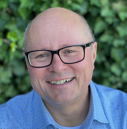 Ein professionelles Porträtfoto vor einem unscharfen grünen Hintergrund. Die Person trägt eine schwarze Brille und ein hellblaues Hemd mit kleinem Muster. Das Bild strahlt eine freundliche, einladende Atmosphäre aus und scheint in einem geschäftlichen Kontext verwendet zu werden, wie etwa als Mitarbeiterporträt oder Ansprechpartner-Foto.