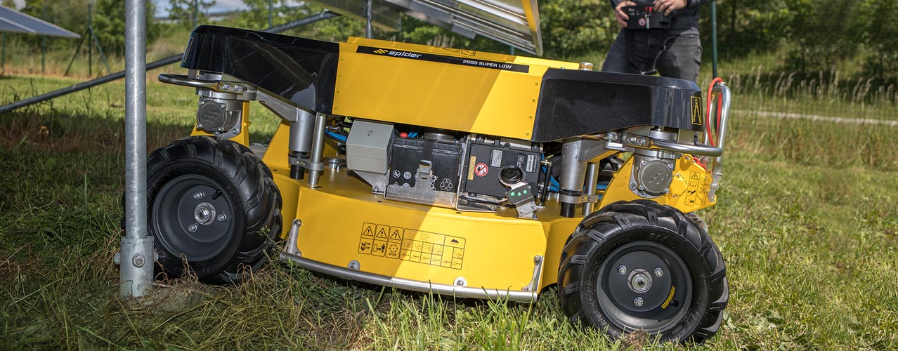 Auf diesem Bild ist der Spider 2SGS EFI Super Low zu sehen, der unter einer Solarpanel-Anlage hindurchfährt. Daneben steht die Bedienperson mit einer Fernbedienung.